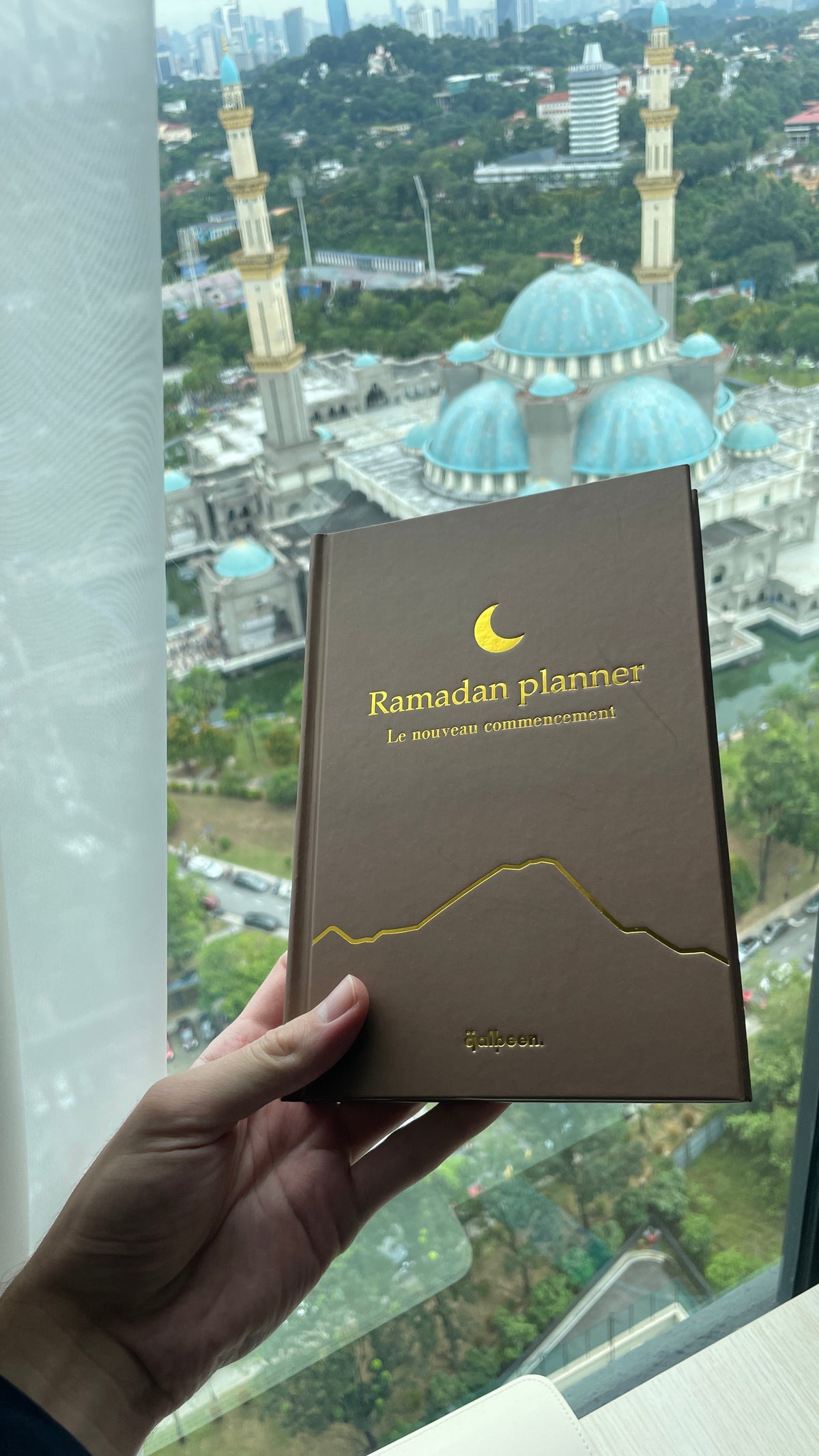 The Ramadan Planner held up in front of a mosque